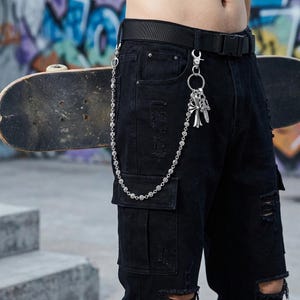 May include: Black cargo pants with distressed details and a silver chain wallet accessory. The chain features a decorative cross pendant and is attached to the belt loop. A skateboard is visible in the background.