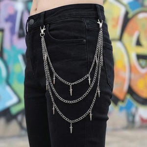 May include: Silver-colored chain accessory with three layers of chains, each adorned with small dagger charms. The chains are attached to the black denim pants with silver clasps. The background features a colorful graffiti wall.