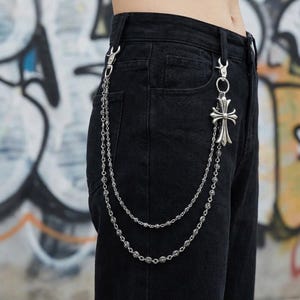 Op de afbeelding: Zilverkleurige kettingportemonnee-accessoire met een decoratieve kruisvormige hanger. De ketting is bevestigd aan de riemlussen van een zwarte denim jeans. De kruishanger is een opvallend kenmerk.