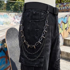 May include: Black denim trousers with distressed detailing and a black belt. A silver chain with cone-shaped spikes hangs from the belt loop. The trousers have a cargo pocket and are worn with a black belt.