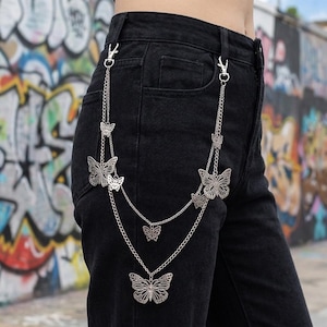 May include: Silver-colored butterfly chain accessory attached to black denim pants. The chain features multiple butterfly pendants of varying sizes, connected by delicate chains and clasps. The background shows a colorful graffiti wall.