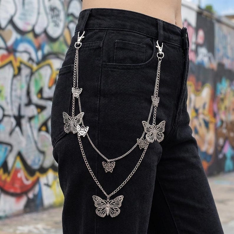May include: Silver-colored butterfly chain accessory attached to black denim pants. The chain features multiple butterfly pendants of varying sizes, connected by delicate chains and clasps. The background shows a colorful graffiti wall.