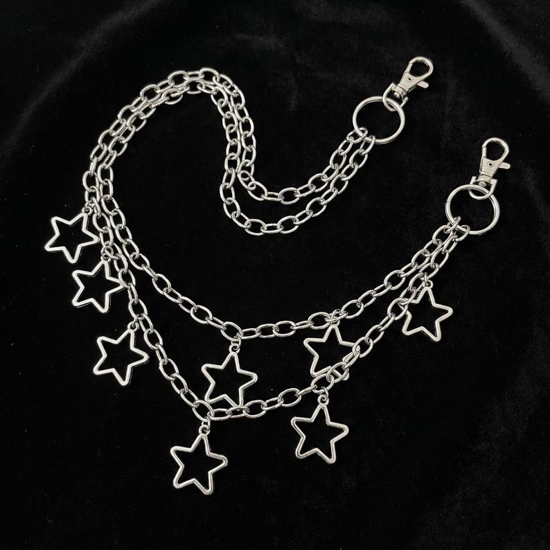 May include: Silver-toned chain belt with multiple strands and star-shaped charms. The belt features two lobster claw clasps and is designed to be worn around the waist or hips. The chains and charms are made of a shiny, metallic material.