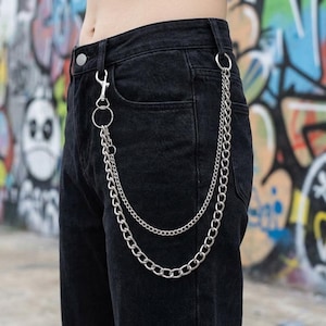 May include: Close-up of black denim pants with a silver chain accessory. The chain has two strands and is attached to the belt loops and a front pocket. The background features colorful graffiti art.
