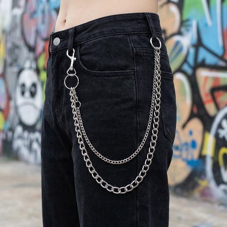 May include: Close-up of black denim pants with a silver chain accessory. The chain has two strands and is attached to the belt loops and a front pocket. The background features colorful graffiti art.
