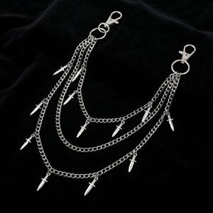 May include: Silver-colored chain accessory with multiple layers and small dagger charms. The accessory features two clasps for attachment and is displayed against a black background. The chains are connected by small rings.