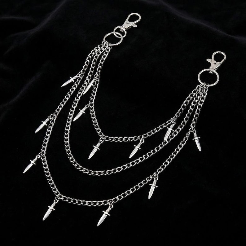 May include: Silver-colored chain accessory with multiple layers and small dagger charms. The accessory features two clasps for attachment and is displayed against a black background. The chains are connected by small rings.
