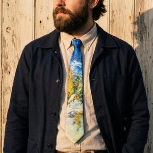 Puede incluir: Una corbata azul con un diseño de pintura de paisaje en tonos azules, amarillos y verdes. La corbata se lleva con una camisa clara y una chaqueta azul marino. El diseño de la corbata recuerda a una obra de arte famosa.