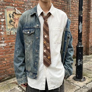 May include: A denim jacket with a light wash, worn over a white button-down shirt and a brown tie. The tie features a pattern of stars and planets. The shirt has a distressed look.