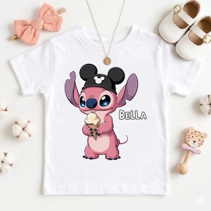 May include: White t-shirt featuring a pink cartoon character wearing a black hat with Mickey Mouse ears and holding an ice cream cone. The word "BELLA" is printed on the shirt. A pink bow and white shoes are also visible.