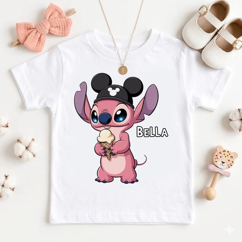 May include: White t-shirt featuring a pink cartoon character wearing a black hat with Mickey Mouse ears and holding an ice cream cone. The word "BELLA" is printed on the shirt. A pink bow and white shoes are also visible.