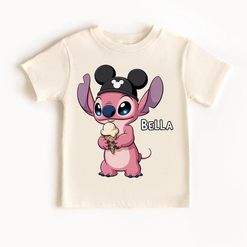May include: Cream-colored t-shirt featuring a pink cartoon character wearing a black hat with Mickey Mouse ears and holding an ice cream cone. The word "BELLA" is printed in black on the shirt.