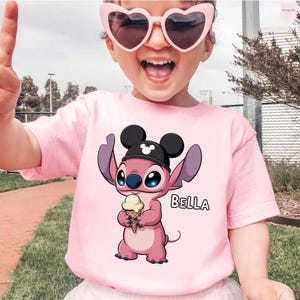 May include: Pink t-shirt featuring a cartoon character holding an ice cream cone, wearing a black hat with Mickey Mouse ears. The name "BELLA" is printed in white. The child is wearing pink heart-shaped sunglasses.