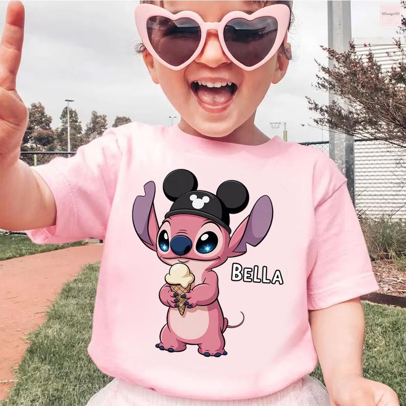 May include: Pink t-shirt featuring a cartoon character holding an ice cream cone, wearing a black hat with Mickey Mouse ears. The name "BELLA" is printed in white. The child is wearing pink heart-shaped sunglasses.