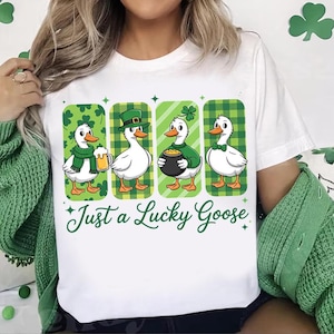 Pode incluir: Camiseta branca com um gráfico de quatro gansos de desenho animado em trajes verdes e brancos do Dia de São Patrício. Os gansos estão em molduras xadrez verdes e brancas e com tema de trevo. O texto "Just a Lucky Goose" está escrito em verde.