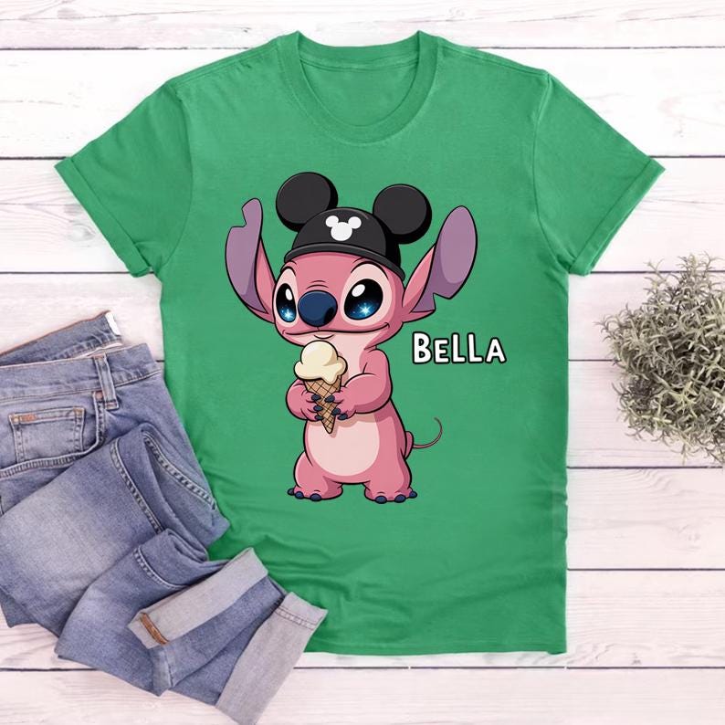 May include: Green t-shirt featuring a pink cartoon character wearing a black hat with Mickey Mouse ears and holding an ice cream cone. The word "BELLA" is printed in white. Blue jeans are visible in the image.