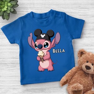 May include: Blue short-sleeved t-shirt featuring a pink cartoon character wearing a black hat with mouse ears and holding an ice cream cone. The name "BELLA" is printed in white. A teddy bear is in the background.