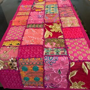 May include: A colourful patchwork table runner in pink, fuchsia, and red hues. The runner is made from various patterned fabrics, including floral and paisley designs, with embroidered details. It has a solid pink border, creating a decorative accent for a table.