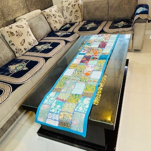 May include: A patchwork table runner with a blue border is displayed on a dark, rectangular coffee table. The runner features a variety of patterned squares in shades of blue, green, yellow, and red. The table is in front of a beige sectional sofa with patterned cushions.