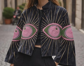 Può includere: Giacca in velluto nero con un design a occhi. La giacca presenta occhi rosa e verdi ricamati con accenti dorati a raggi. Ha un collo alto e maniche svasate. La modella indossa pantaloni neri.