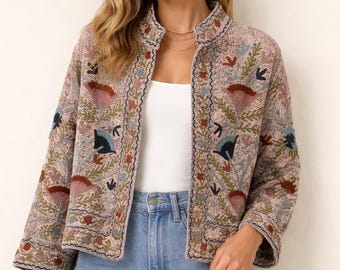 Puede incluir: Chaqueta bordada rosa claro con cuello alzado y mangas tres cuartos. La chaqueta presenta motivos florales y de hojas en tonos azules, marrones y verdes. La chaqueta se combina con vaqueros azul claro.
