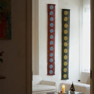 May include: Two vertical wall hangings with a repeating dot pattern. One is a deep red with blue dots, the other is dark green with yellow dots. Both are mounted on the wall with wooden dowels. The room has white furniture and a lit candle.