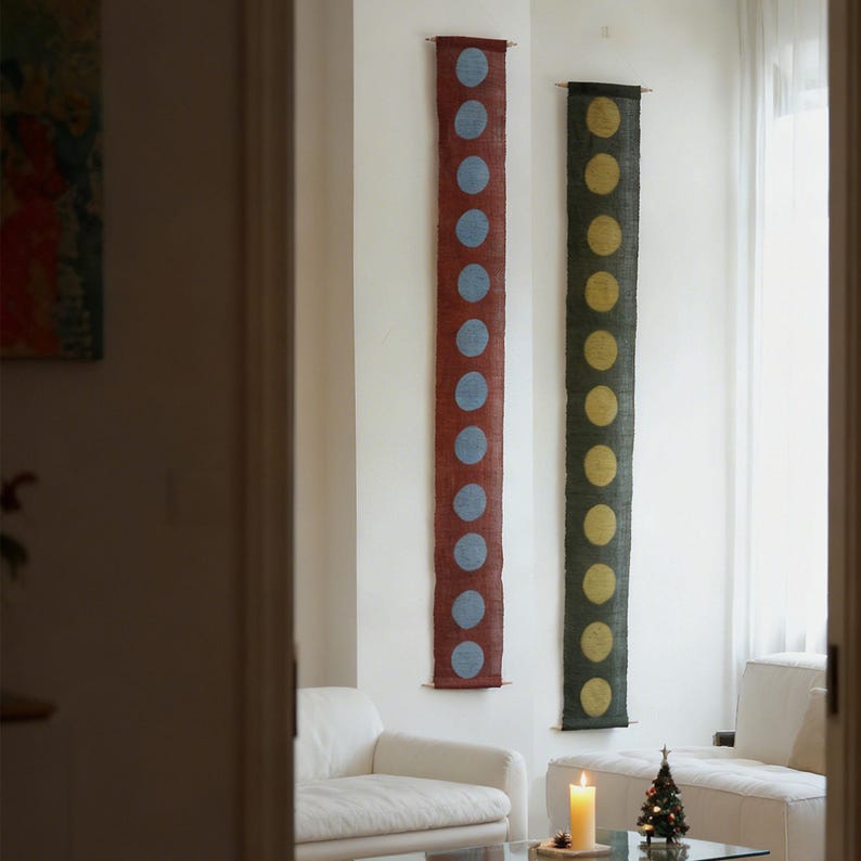 May include: Two vertical wall hangings with a repeating dot pattern. One is a deep red with blue dots, the other is dark green with yellow dots. Both are mounted on the wall with wooden dowels. The room has white furniture and a lit candle.