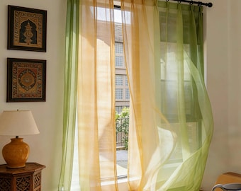 May include: Sheer curtains in green and yellow-orange hang from a black rod. The light filters through the fabric, creating a soft glow. The curtains are layered, adding depth to the window. Decorative framed art and a lamp are visible. The curtains are approximately 200 cm in length.