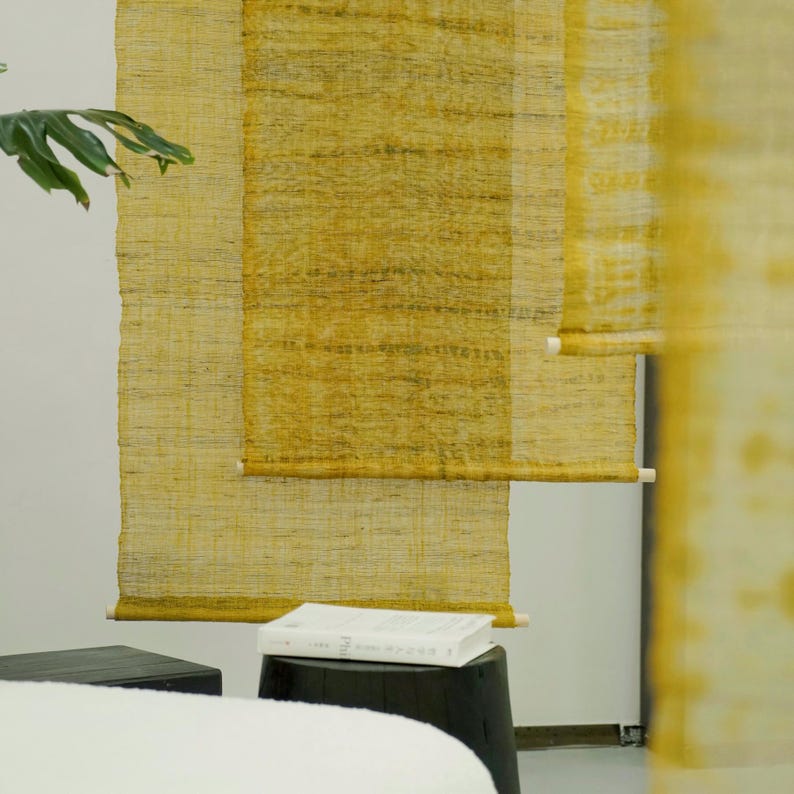 May include: Yellow woven window shades hang against a white wall. The shades are made of a textured material and have a natural, organic look. A white book rests on a black stool in front of the shades. A green plant is visible on the left.