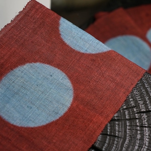 May include: Close-up of a red fabric featuring large, light blue polka dots. The fabric has a textured appearance and is draped over a dark, wooden surface. The polka dot pattern is repeated across the fabric.