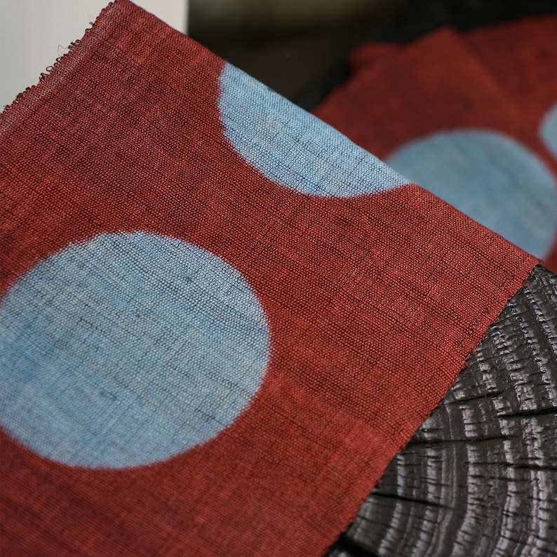 May include: Close-up of a red fabric featuring large, light blue polka dots. The fabric has a textured appearance and is draped over a dark, wooden surface. The polka dot pattern is repeated across the fabric.