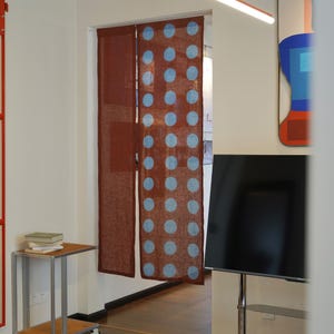 May include: A pair of brown linen curtains with a pattern of large, light blue polka dots. The curtains hang in a doorway, with a glimpse of a television and artwork visible in the background. The curtains add a decorative touch to the room.