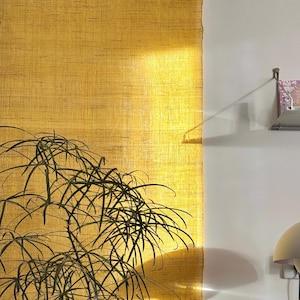May include: A textured, golden-yellow wall hanging with a natural, woven appearance. The sunlight casts a warm glow across the fabric. A green plant with long, thin leaves is in the foreground. A small shelf with books is visible on the right.