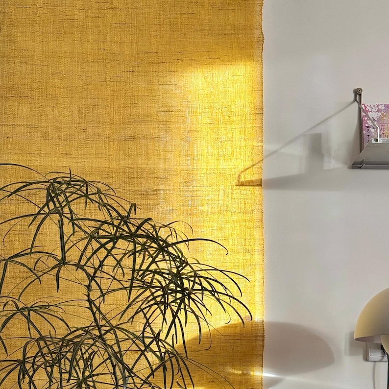 May include: A textured, golden-yellow wall hanging with a natural, woven appearance. The sunlight casts a warm glow across the fabric. A green plant with long, thin leaves is in the foreground. A small shelf with books is visible on the right.