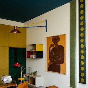 May include: Interior design with a modern aesthetic. A copper-coloured wall lamp is mounted on the wall. A framed painting of a woman hangs on the wall. A red desk lamp sits on a glass desk. The colour scheme is yellow and green.
