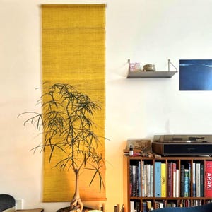 May include: A tall, vertical, yellow woven wall hanging is mounted on a white wall. A small tree with thin, green leaves is in front of the wall hanging. A bookshelf with books and a record player are in the background.