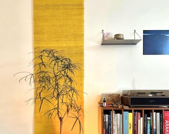 May include: A long, vertical, yellow woven wall hanging is mounted on a white wall. A small tree with thin, green leaves is in front of the wall hanging. A bookshelf with books and a record player are in the background.
