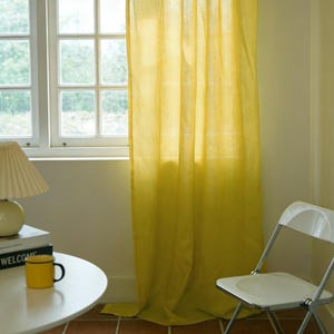 Pode incluir: Uma cortina amarela brilhante está pendurada ao lado de uma janela branca. Uma mesa branca tem uma lâmpada e uma caneca amarela. Uma cadeira dobrável branca está por perto. O quarto tem uma atmosfera acolhedora.