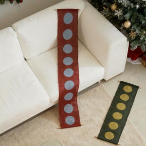 May include: Two decorative table runners. One is a deep red with large, light blue polka dots. The other is dark green with yellow polka dots. Both have wooden dowels at each end. The runners are displayed on a white sofa and a beige rug.