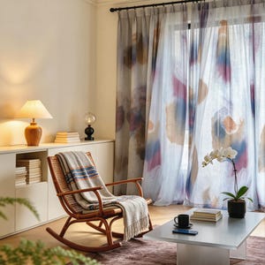 May include: A cozy living room scene featuring a wooden rocking chair with a blanket, a table with a coffee cup, and sheer curtains with colorful abstract patterns. A lamp and a potted orchid add to the inviting atmosphere. The room has a neutral color palette.