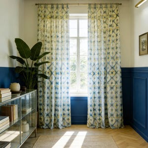 Op de afbeelding: Interieur met gordijnen met blauwe en gele patronen voor een raam. Links staat een grote groene plant. De muren zijn wit en blauw geschilderd, en de vloer is van hout.