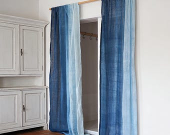 May include: Two blue ombre curtains hanging from a wooden rod. The curtains transition from a deep navy to a light sky blue. The curtains are in front of a doorway, and the room has a white cabinet and terracotta tile flooring.