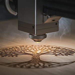 May include: A laser engraving machine etching a detailed tree of life design onto a wooden surface. The machine's nozzle emits a bright light, creating a smoky effect. The wood has a light brown color.