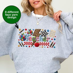 Puede incluir: Una sudadera gris claro de cuello redondo con un gr&aacute;fico colorido que muestra la palabra Teacher en un estilo de hilo falso texturizado. El dise&ntilde;o incluye un rayo con estampado de leopardo, manzanas, l&aacute;pices de colores, estrellas y un patr&oacute;n de cuadros. Un c&iacute;rculo verde con el texto 8 different faux yarn design se&ntilde;ala el gr&aacute;fico.