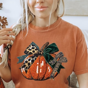 May include: A rust-coloured short-sleeve t-shirt featuring a graphic of a textured orange pumpkin adorned with a leopard print bow and a dark green velvet bow. A banner wrapped around the pumpkin displays the text EST. 1993 PUMPKIN SPICE SEASON.