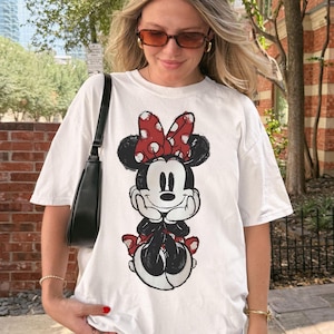 May include: White t-shirt featuring a Minnie Mouse graphic. The cartoon character is depicted in black, red, and white, with a large red polka-dot bow. The shirt is a casual style, suitable for everyday wear.