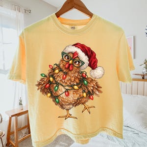 May include: A butter yellow short-sleeve t-shirt featuring a graphic of a brown chicken wearing a red and white Santa hat and green glasses, wrapped in colorful Christmas lights. The Comfort Colors brand logo is printed on the inner neck label.