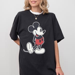 May include: Black t-shirt featuring a large graphic of Mickey Mouse in his classic pose. The cartoon character is depicted in black, red, and white. The shirt has a white collar and short sleeves.