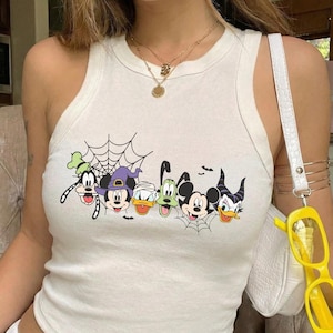 May include: A white sleeveless tank top featuring a graphic print of various cartoon characters including Mickey Mouse, Minnie Mouse, Donald Duck, and Goofy in Halloween costumes, accented with spider webs and small bats across the chest area.