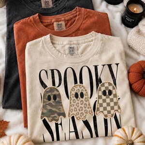May include: Three crew neck t-shirts in black, rust orange, and cream. The cream shirt features the text SPOOKY SEASON in black with three ghost graphics, each with a different pattern: camouflage, leopard print, and a tan and white checkerboard.